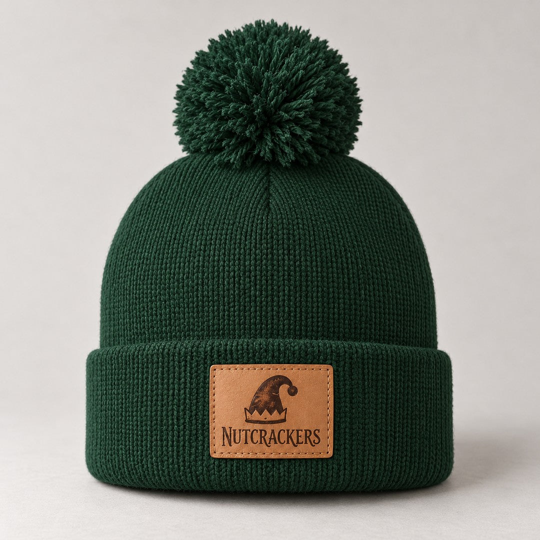 Forest-green pom-pom beanie with a leather patch engraved with an elf hat and the word Nutcrackers