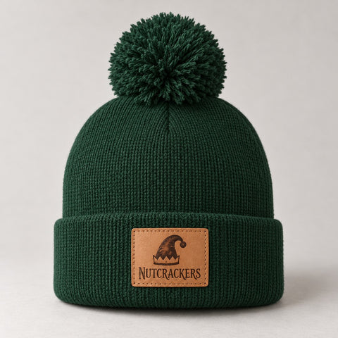 Forest-green pom-pom beanie with a leather patch engraved with an elf hat and the word Nutcrackers