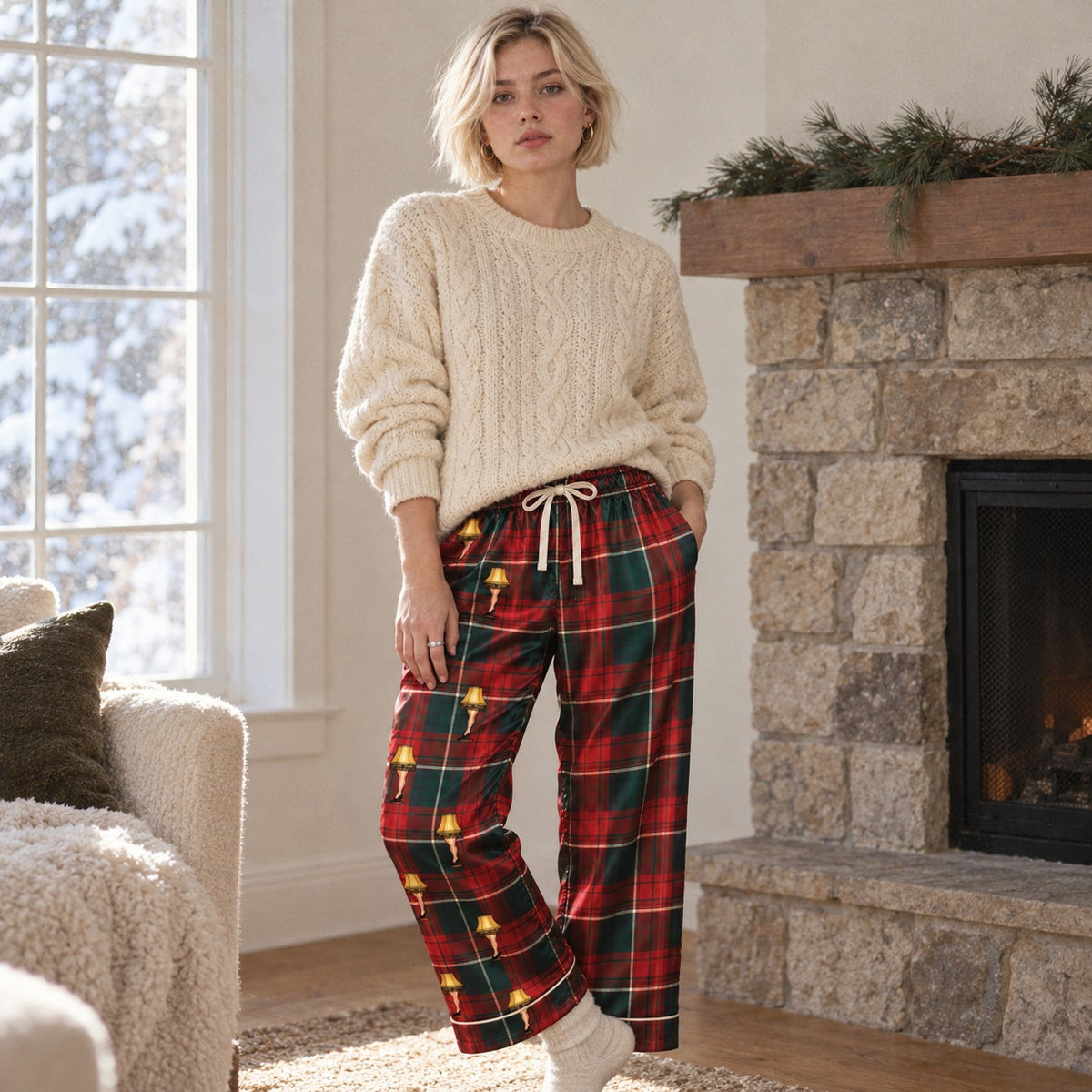 Woman in a cream sweater wearing tartan satin pajama pants by a stone fireplace